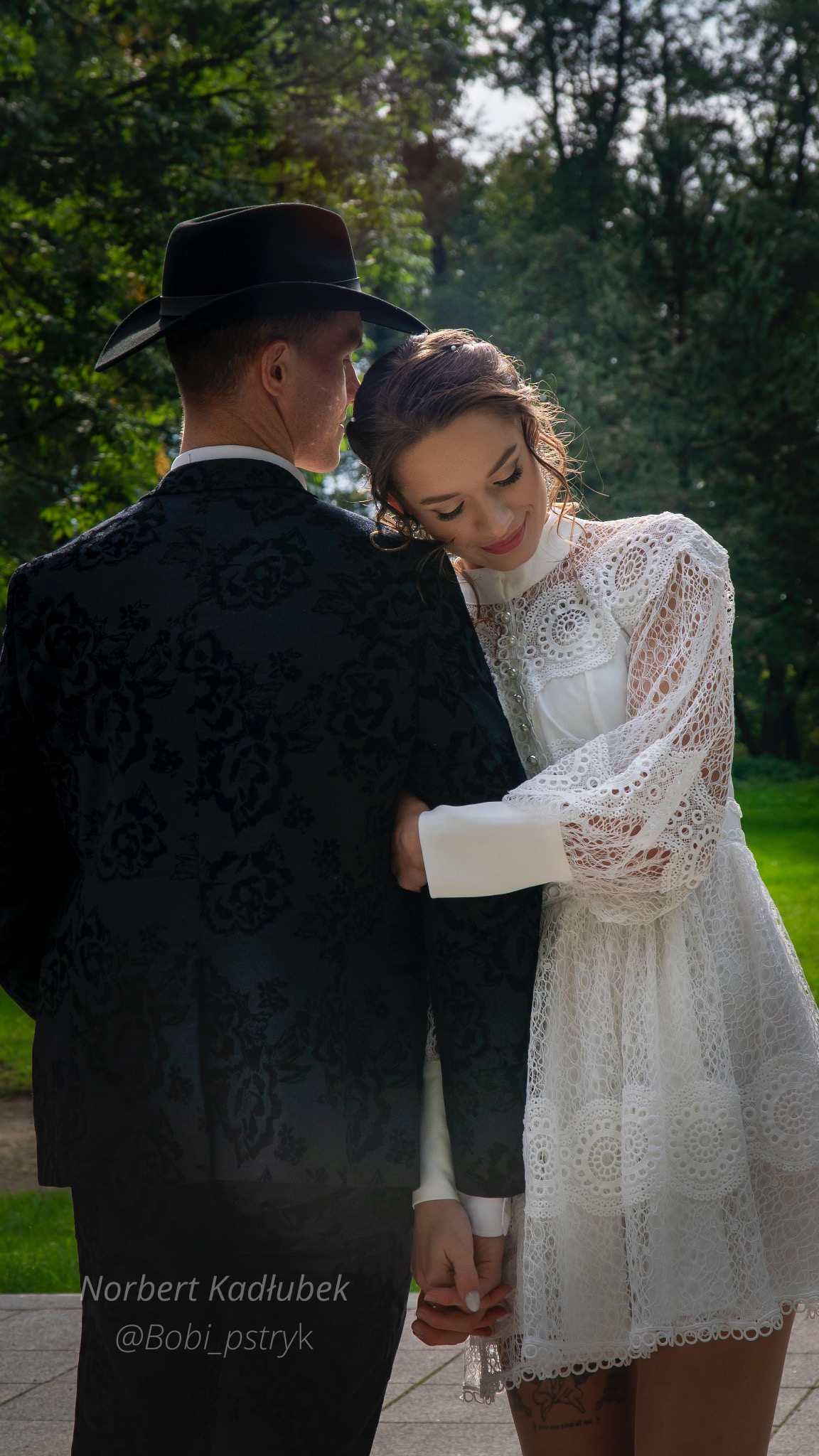 Panna młoda w koronkowej sukni opiera głowę na ramieniu pana młodego w czarnym garniturze z kwiatowym wzorem i kapeluszu, trzymając się za ręce na tle zieleni.