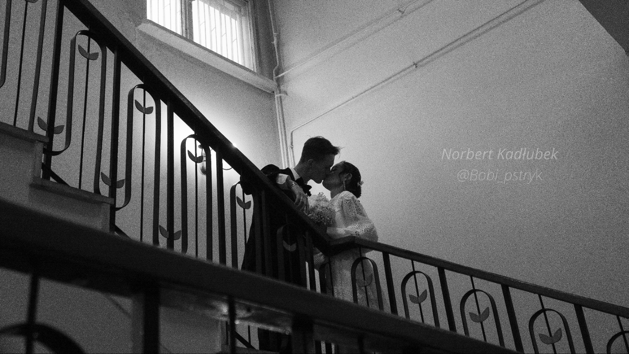 Czarno-białe zdjęcie nowożeńców całujących się na schodach z metalową balustradą, panna młoda w koronkowej sukni trzyma bukiet, widok z dołu.