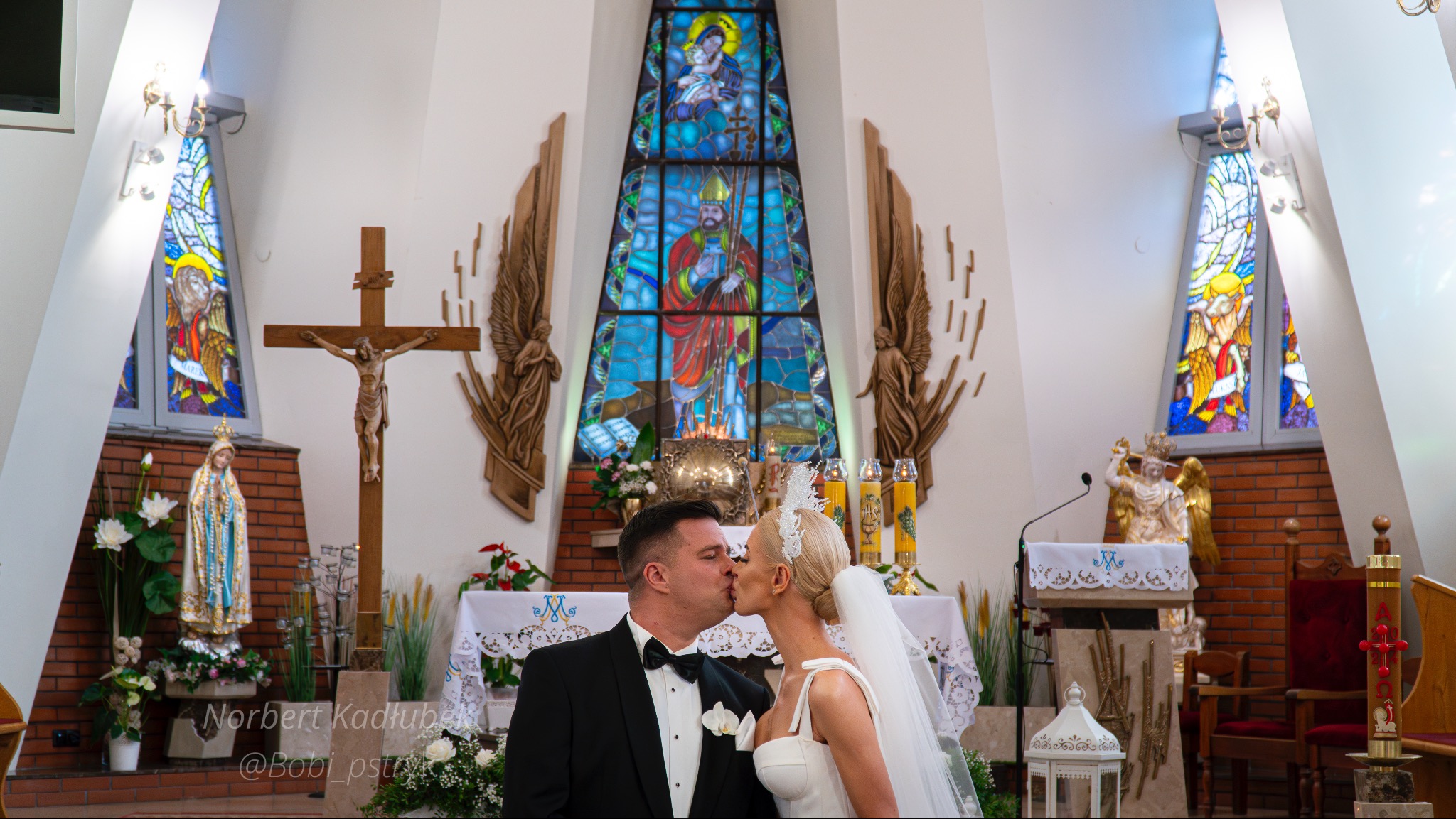 Para młoda całuje się przed ołtarzem w kościele, na tle witraży i dekoracji.