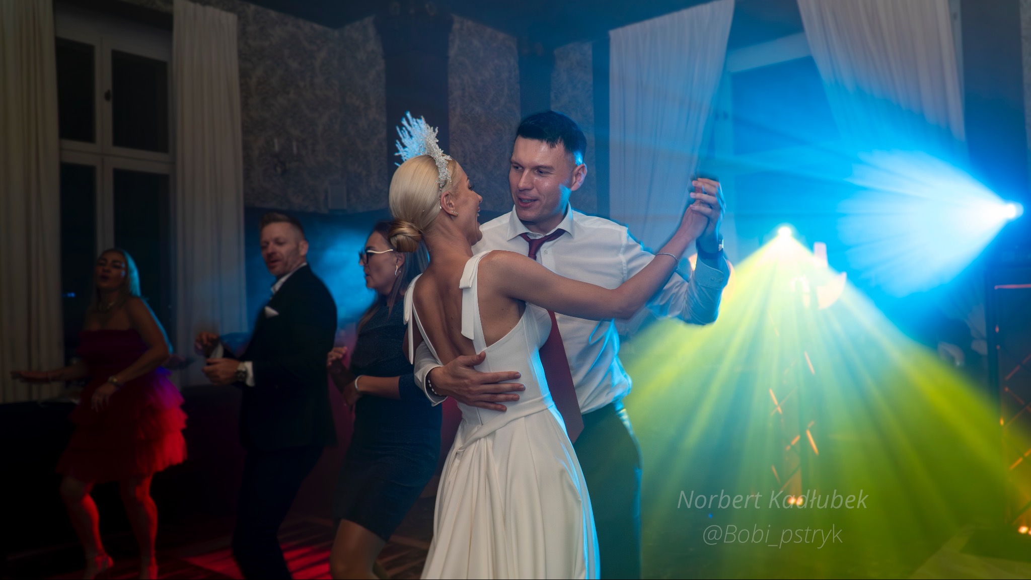 Para młoda tańczy w objęciach na sali weselnej, oświetlona kolorowymi światłami, w tle bawiący się goście.