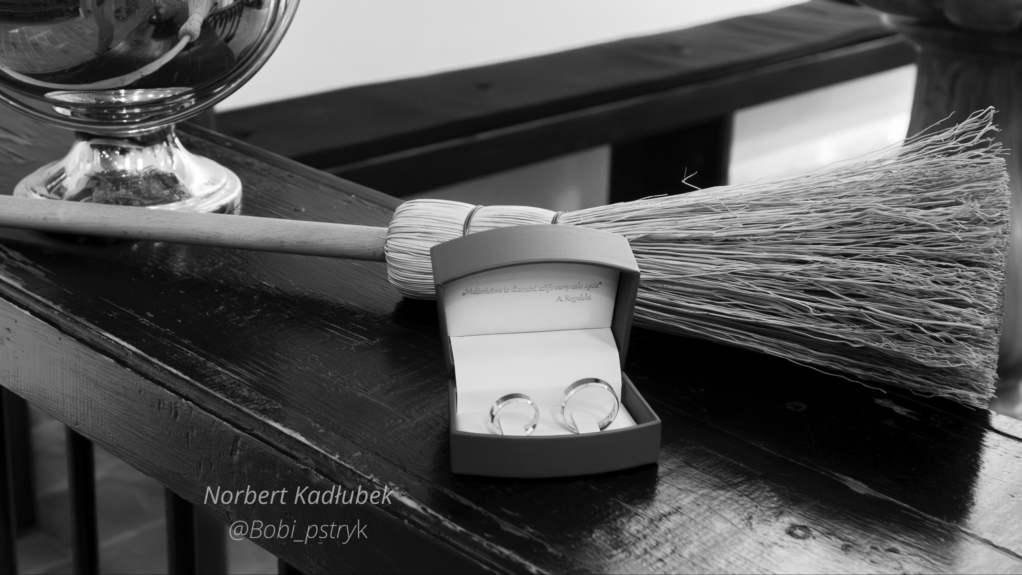 Czarno-białe ujęcie dwóch obrączek ślubnych w pudełku z cytatem, leżących obok miotły na lakierowanym drewnianym stole, z odbiciem w szklanej misie w tle.
