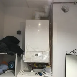 Nowy, biały piec gazowy De Dietrich, jeszcze w folii ochronnej, zamontowany na ścianie w pomieszczeniu. Widoczne rury miedziane, wentylacja, gniazdko elektryczne, półka z ubraniami i narzędzia.