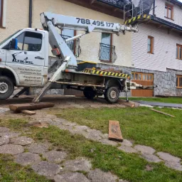 Pracownik na podnośniku koszowym, zabezpieczający dach budynku obłożony czarną folią, z numerem telefonu firmy na wysięgniku i logo firmy Alpin-Mal na samochodzie.