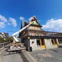 Pracownik na podnośniku koszowym czyści dach i drewniane elementy dekoracyjne budynku z napisem 'Panorama', widoczne ślady mycia wodą pod ciśnieniem.