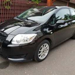 Błyszczący, czarny samochód Toyota Auris zaparkowany na asfalcie, widoczny pod kątem z przodu i z boku, z aluminiowymi felgami i odbiciami w lakierze.
