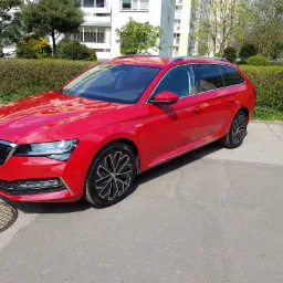 Czerwona Skoda Superb Combi zaparkowana w Łodzi na tle zieleni i budynku mieszkalnego. Widoczny fragment studzienki kanalizacyjnej obok przedniego zderzaka.