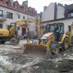 Dwie żółte maszyny budowlane CAT, koparka kołowa M313C i koparko-ładowarka 428F, pracują na terenie budowy z gruzem i ziemią, na tle budynków mieszkalnych.