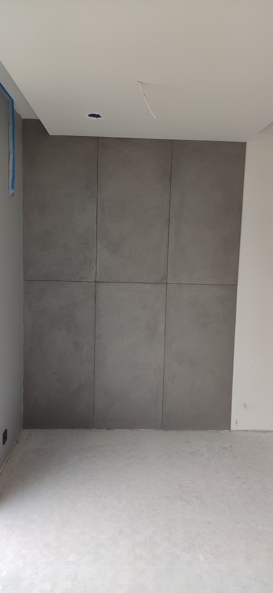 Ściana wyłożona sześcioma szarymi płytami imitującymi beton, widoczne fugi, surowy styl, gładka powierzchnia podłogi, przygotowania do montażu oświetlenia sufitowego.