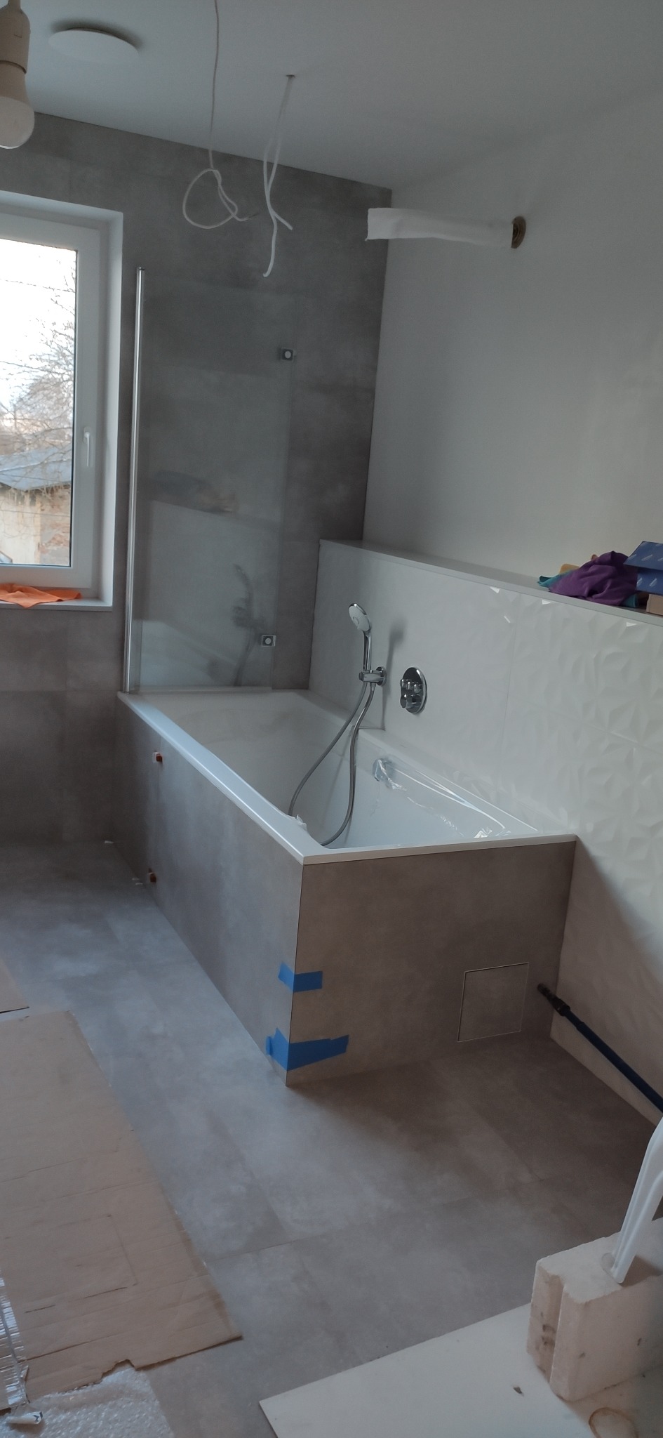 Wanna obudowana płytkami imitującymi beton, z zamontowaną szklaną ścianką prysznicową i armaturą, w trakcie prac wykończeniowych, widoczne zabezpieczenia i niedokończone instalacje.