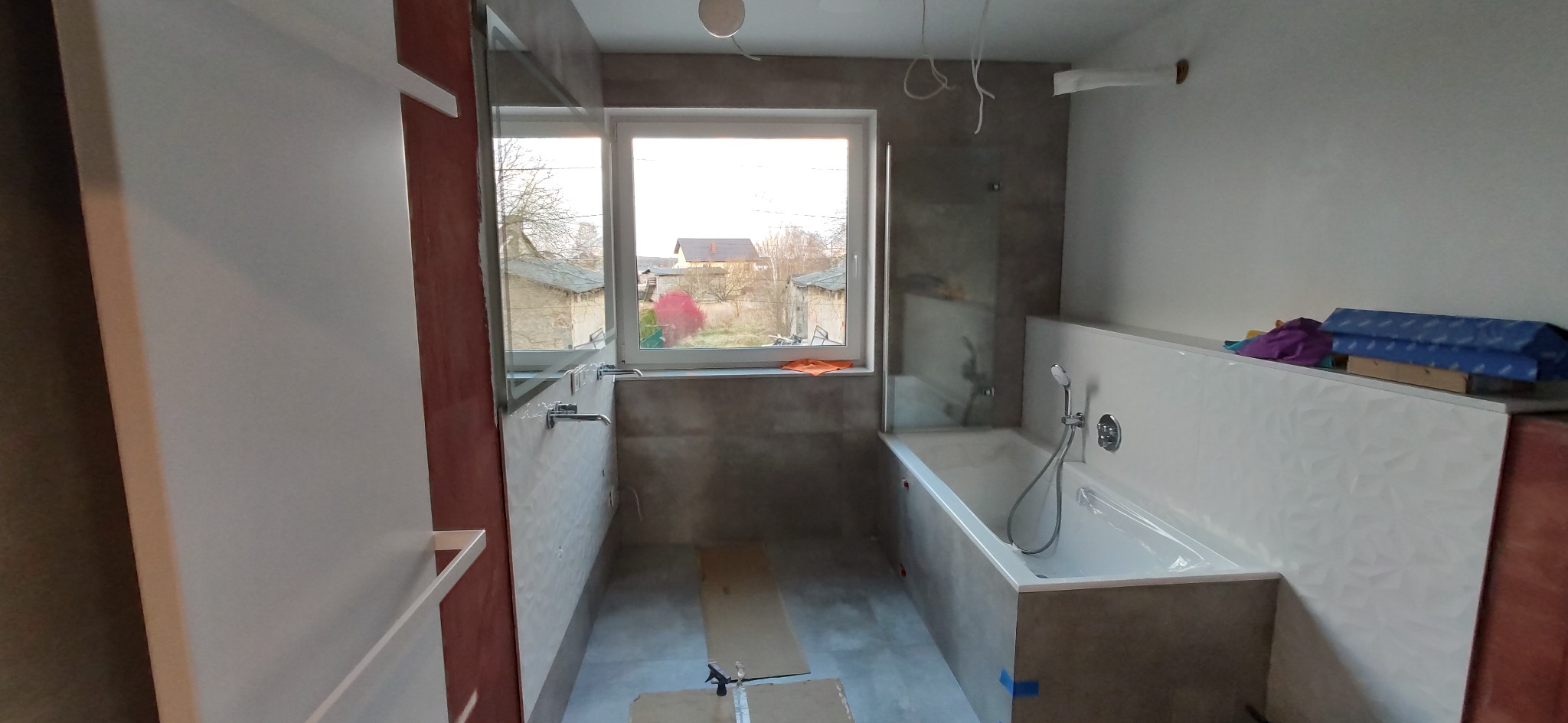 Łazienka w trakcie remontu: wanna obudowana szarą płytką, częściowo przeszklona kabina prysznicowa, okno z widokiem na zabudowania, przygotowane płytki na obudowie wanny, surowy stan ścian.