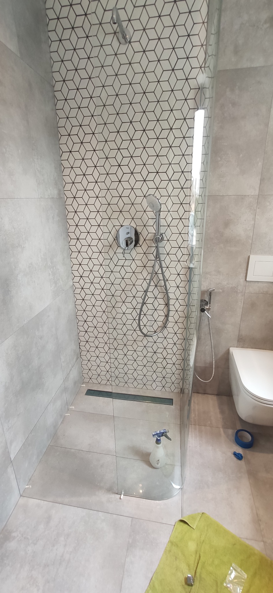 Nowoczesna kabina prysznicowa z geometryczną mozaiką na ścianie, widoczny odpływ liniowy, bateria podtynkowa, bidetka oraz toaleta podwieszana, na podłodze leży zielona ścierka i butelka ze...