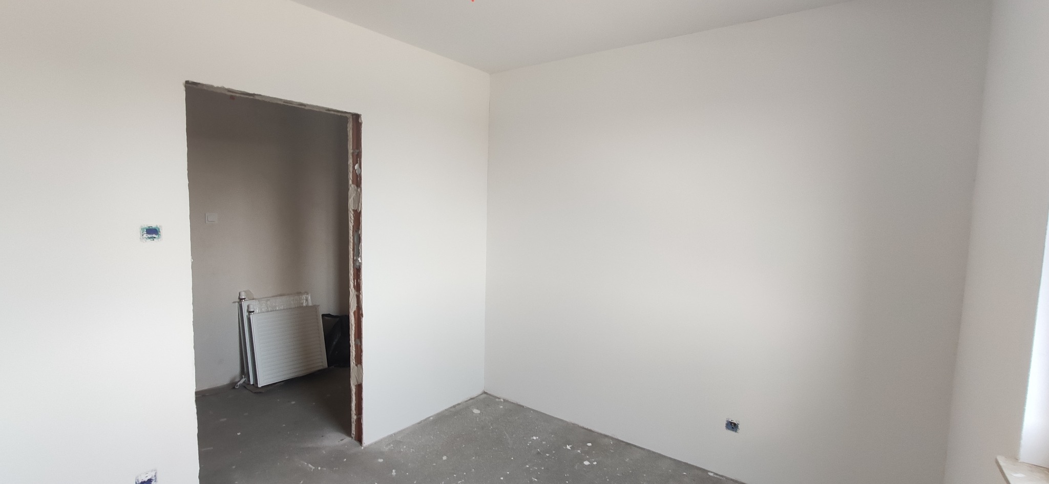 Wnętrze w trakcie remontu: białe ściany po malowaniu, widoczne gniazdka elektryczne, surowa ościeżnica drzwiowa, grzejnik w folii w sąsiednim pomieszczeniu, szary beton na podłodze z resztkami gruzu.