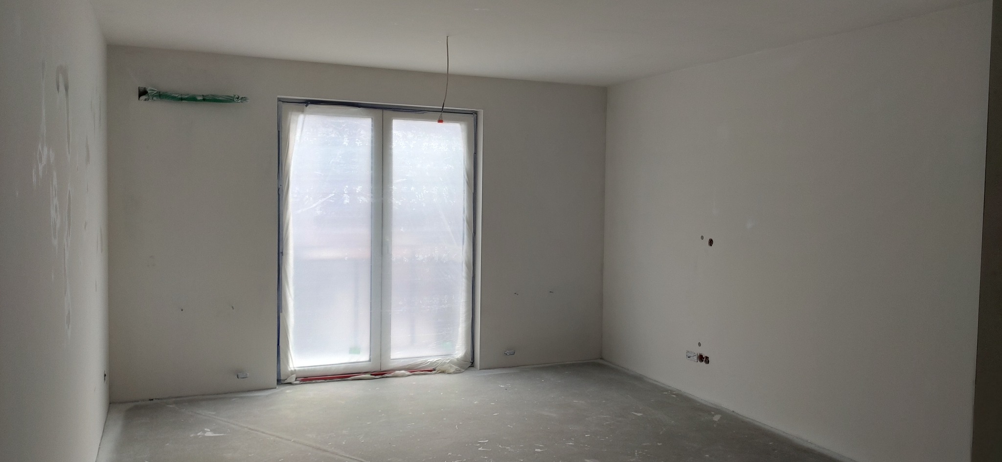 Pusty pokój po szpachlowaniu ścian, okno zabezpieczone folią, widoczne puszki elektryczne i instalacja pod klimatyzację.