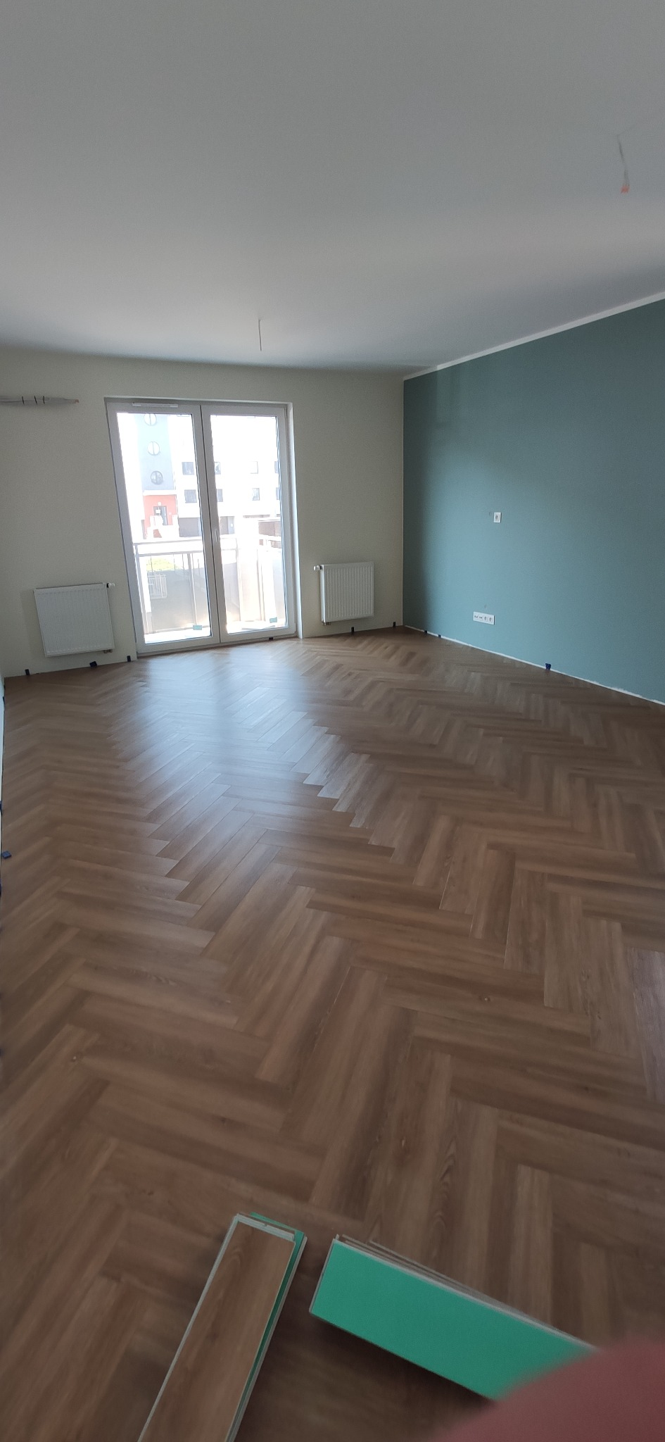 Ukończony pokój z parkietem ułożonym we wzór jodełki, pomalowana na niebiesko ściana, białe ściany z grzejnikami, okno balkonowe z widokiem na budynek mieszkalny, paczki paneli podłogowych w rogu...