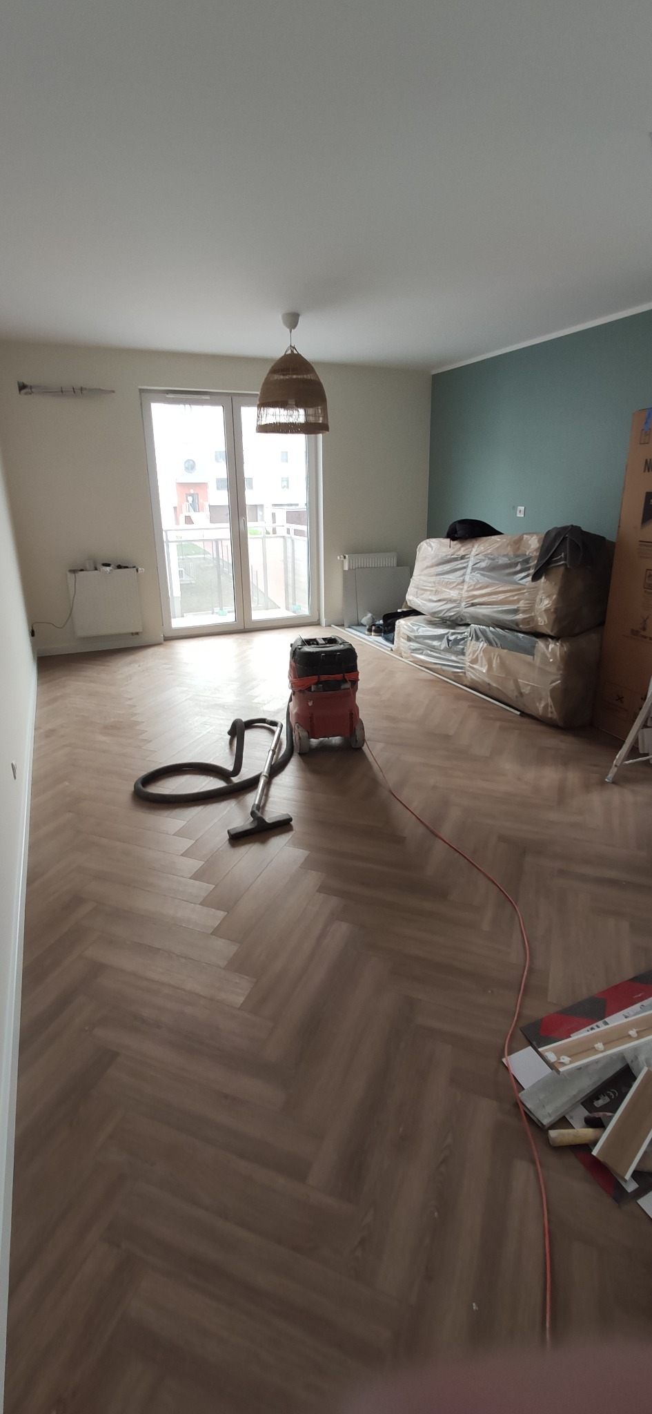 Ułożona podłoga w jodełkę, odkurzacz przemysłowy na środku pokoju, w tle okno balkonowe i zapakowane meble.