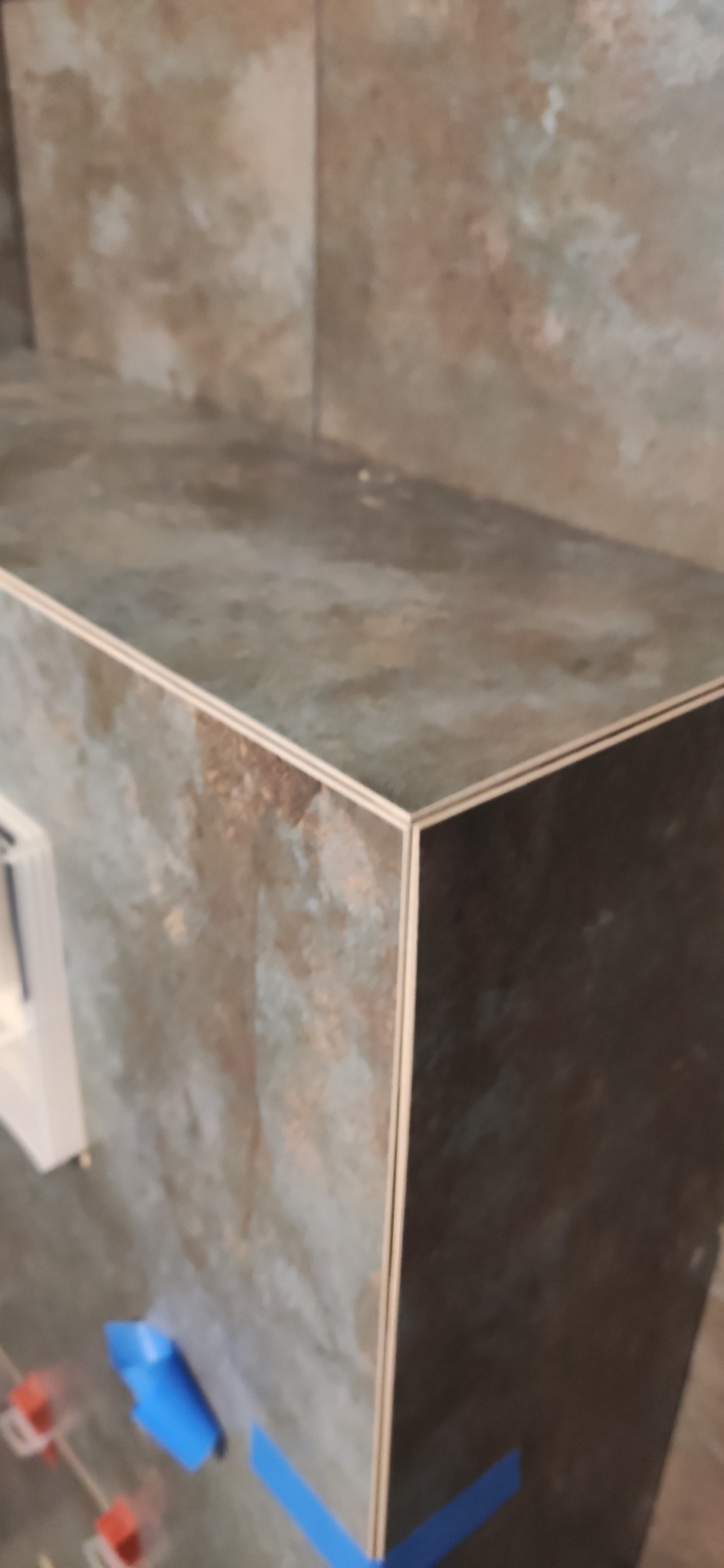 Wykończona obudowa z płytek imitujących kamień, zabezpieczona niebieską taśmą malarską i poziomowana za pomocą czerwonych klinów dystansowych.