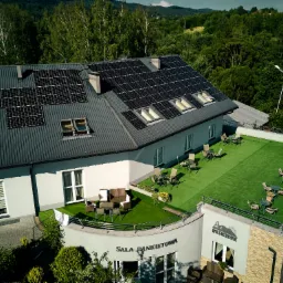 Widok z góry na budynek z panelami słonecznymi na dachu i tarasem z meblami ogrodowymi, otoczony zielenią i górami w tle.