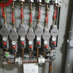 Rozdzielacz instalacji grzewczej z manometrami i zaworami termostatycznymi, widoczne miedziane rury i szara ściana z betonową fakturą.