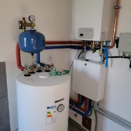 Biały piec gazowy Fondital zawieszony obok pionowego, cylindrycznego zbiornika Kospel z niebieskim naczyniem przeponowym, połączone rurami miedzianymi, czerwonymi i niebieskimi, na tle białej...