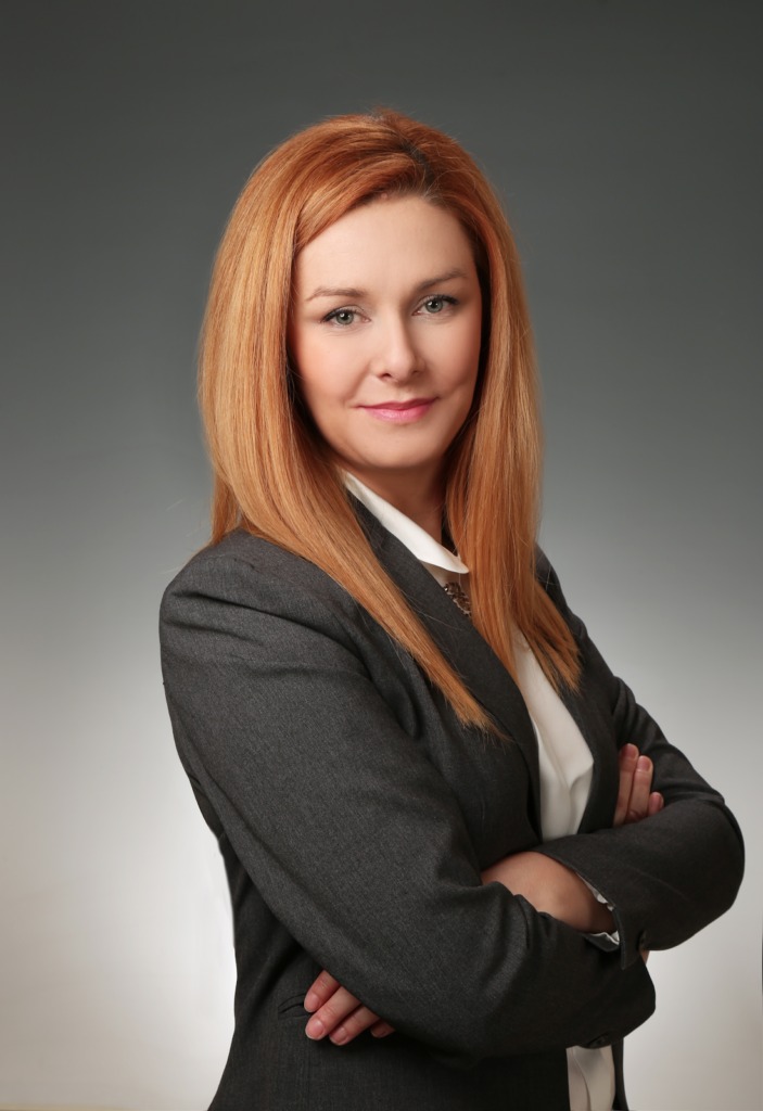 Portret kobiety w szarym żakiecie i białej koszuli, z rudymi włosami, skrzyżowane ramiona, na szarym tle.