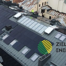 Zielona Energia Sp. z o.o. - Widok z góry na dach kamienicy z zamontowanymi panelami słonecznymi, lukarnami i oknami dachowymi, w tle inne budynki i pracownik na dachu.