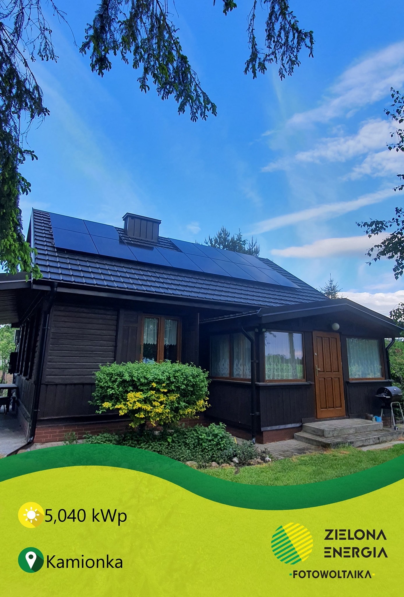 Dom z ciemnego drewna z panelami słonecznymi na dachu, widok z dołu, błękitne niebo z rzadkimi chmurami, zielony trawnik i krzewy.