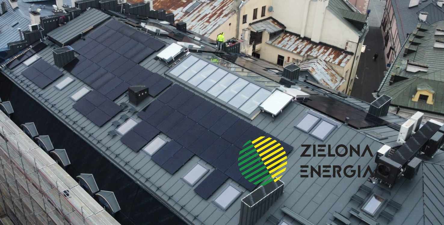Widok z góry na dach kamienicy z zamontowanymi panelami słonecznymi, lukarnami i oknami dachowymi, w tle inne budynki i pracownik na dachu.