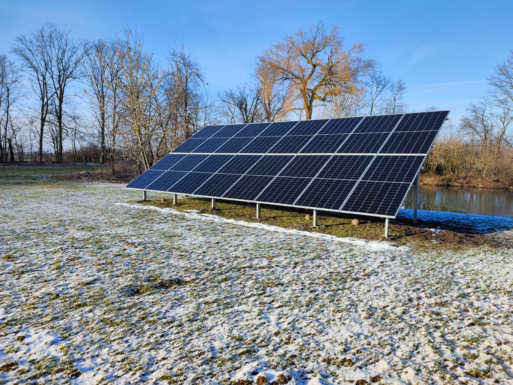 Panel słoneczny ustawiony na trawie pokrytej szronem, w tle nagie drzewa i błękitne niebo.