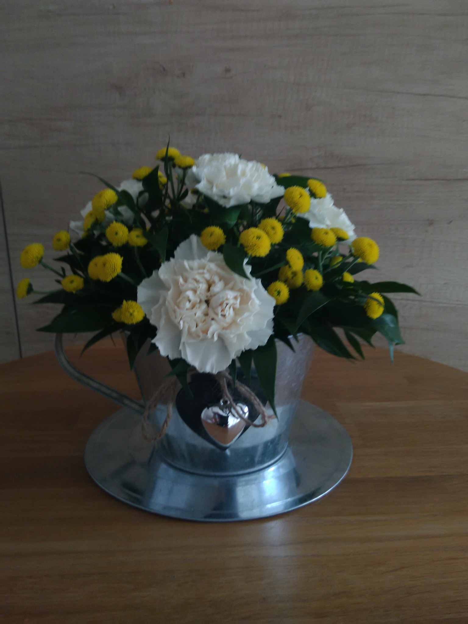 Kompozycja kwiatowa z białych goździków i żółtych chryzantem w metalowym wiaderku z doczepionym srebrnym serduszkiem, Kiełczów. Dekoracja na drewnianym stole.