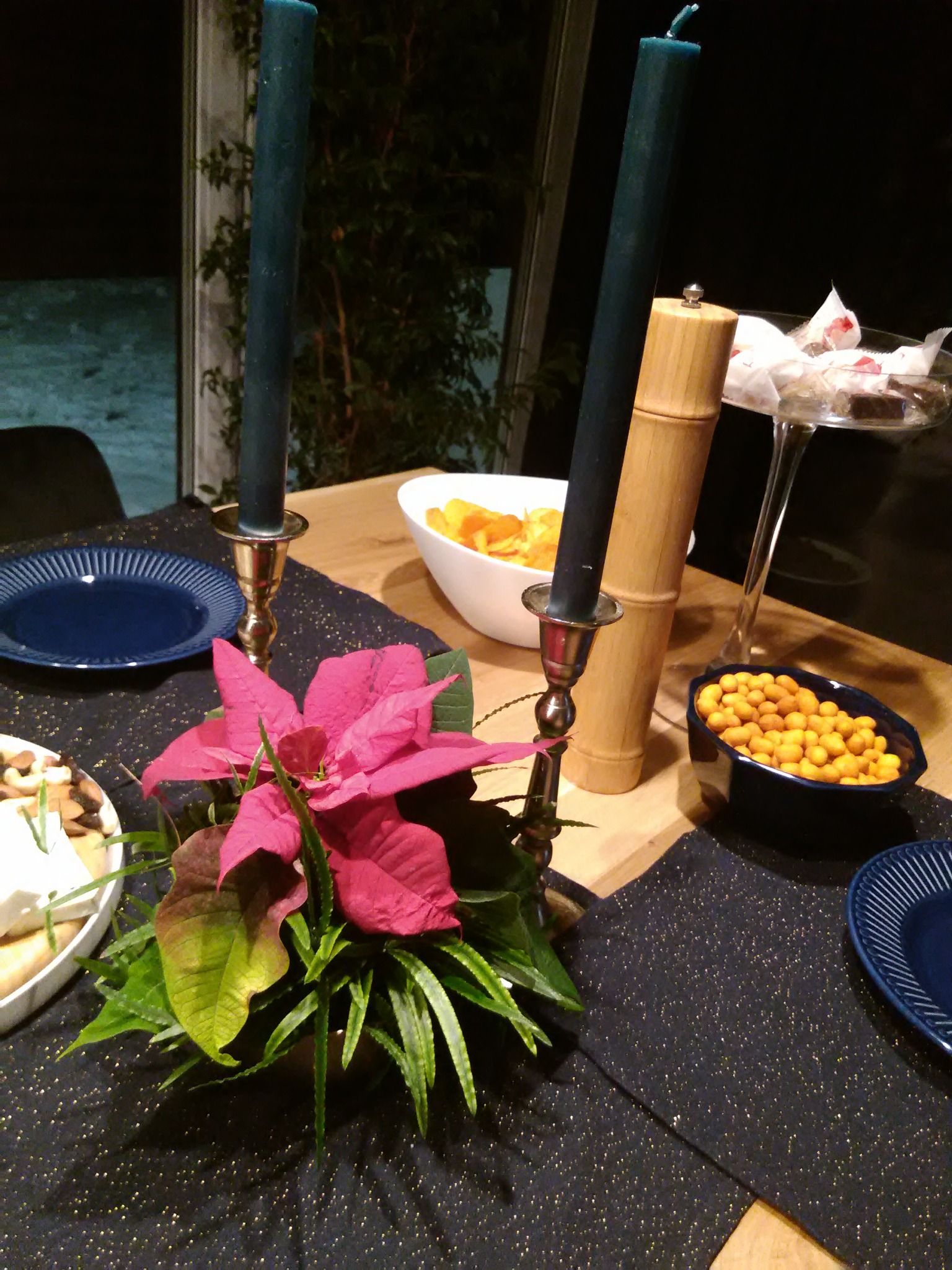 Dekoracja stołu z ciemnoniebieskimi świecami, czerwoną poinsecją, przekąskami i niebieskimi talerzami na błyszczącym, ciemnym bieżniku.