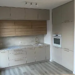 Jasna kuchnia na wymiar z drewnianymi akcentami, złotą armaturą i wbudowanym piekarnikiem, oświetlona trzema reflektorami.