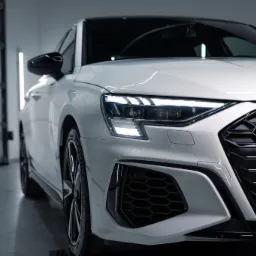 Biały samochód Audi RS3 po umyciu, widok z przodu na reflektory i grill z emblematem RS3, w tle brama garażowa.