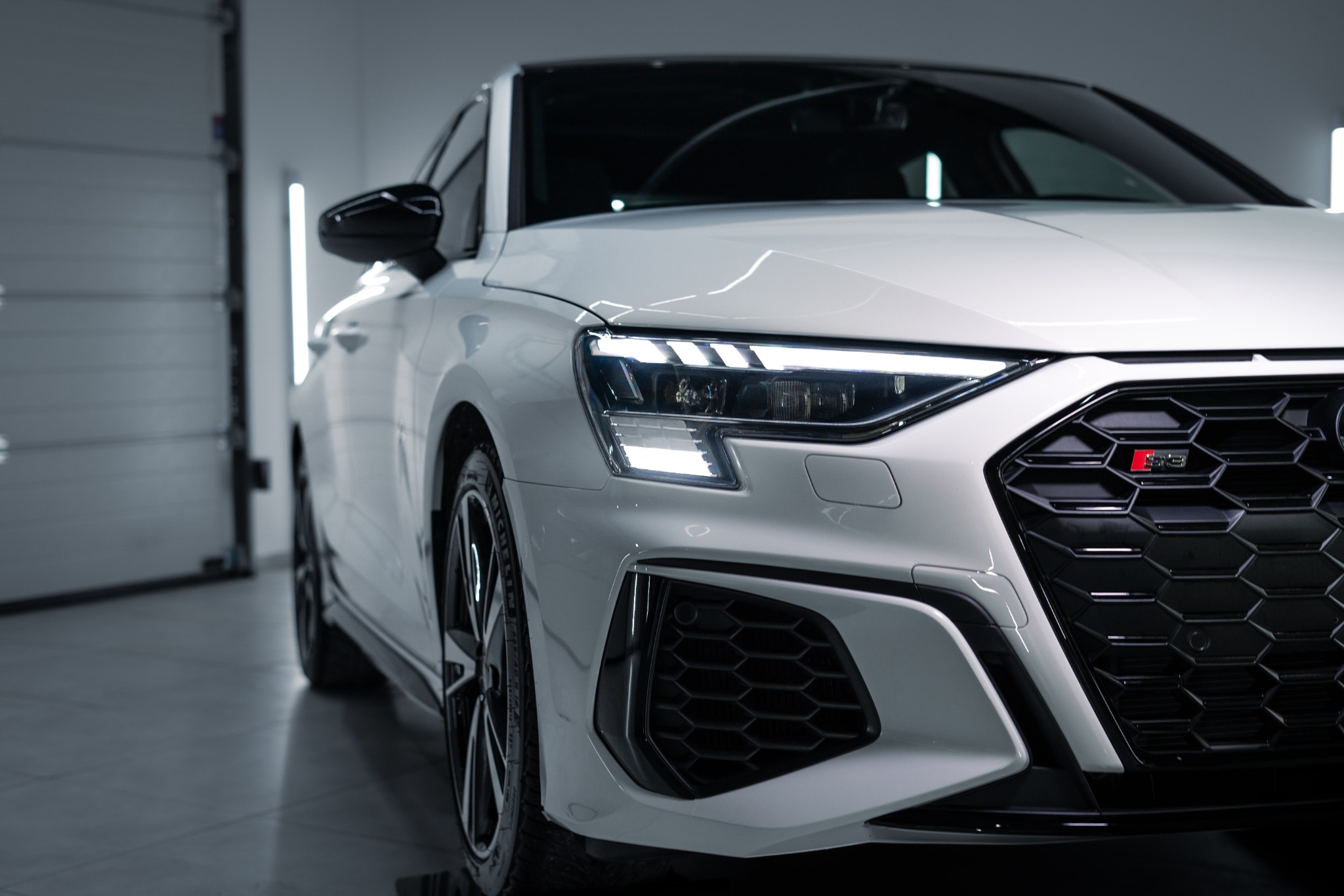 Biały samochód Audi RS3 po umyciu, widok z przodu na reflektory i grill z emblematem RS3, w tle brama garażowa.
