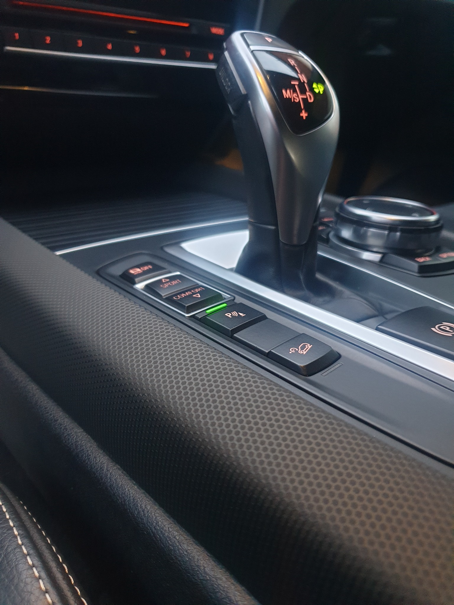 Wnętrze samochodu z bliska: dźwignia zmiany biegów, panel sterowania z podświetlanymi przyciskami (Comfort, Sport, Parking) i fragment deski rozdzielczej z fakturą.
