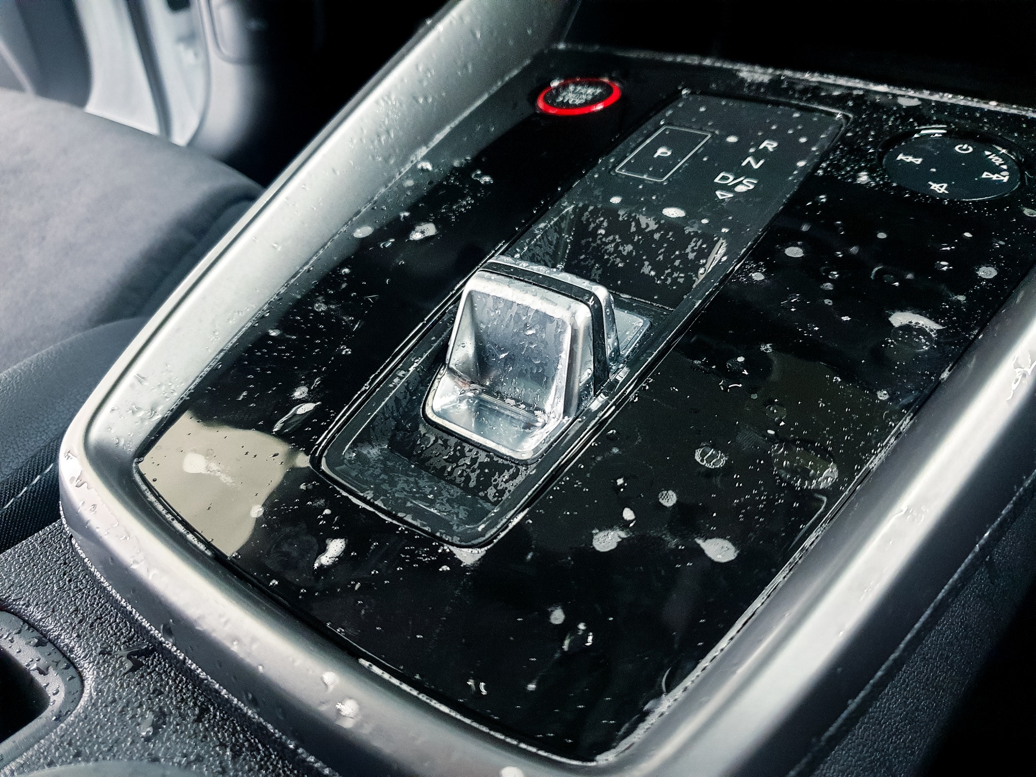 Błyszczący panel zmiany biegów w samochodzie, pokryty kroplami wody lub piany, sugerujący świeże czyszczenie wnętrza pojazdu. Widoczne przyciski i chromowane elementy.
