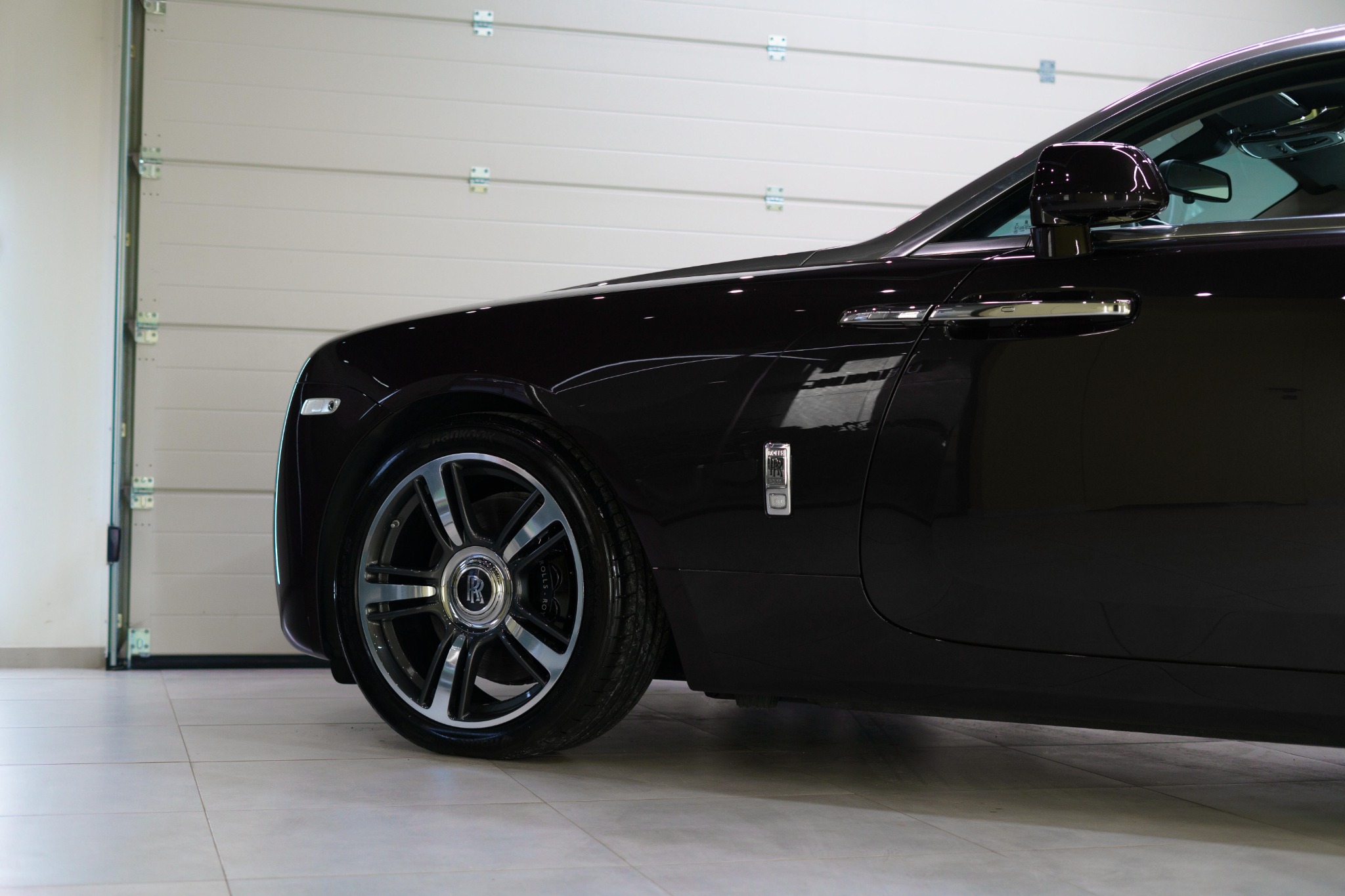 Częściowy widok ciemnobordowego Rolls-Royce'a na tle białej bramy garażowej, z widocznym chromowanym kołem i logiem marki na błotniku.