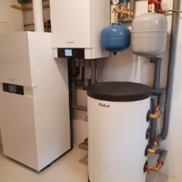 Nowoczesna kotłownia z białym piecem gazowym Viessmann i zasobnikiem Vaillant, rury w szarej izolacji, manometr na instalacji, minimalistyczne wnętrze w jasnych kolorach.