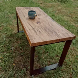 Drewniany stół na metalowych nogach, z widocznym usłojeniem drewna, ustawiony na trawie, z ceramicznym kubkiem na blacie.