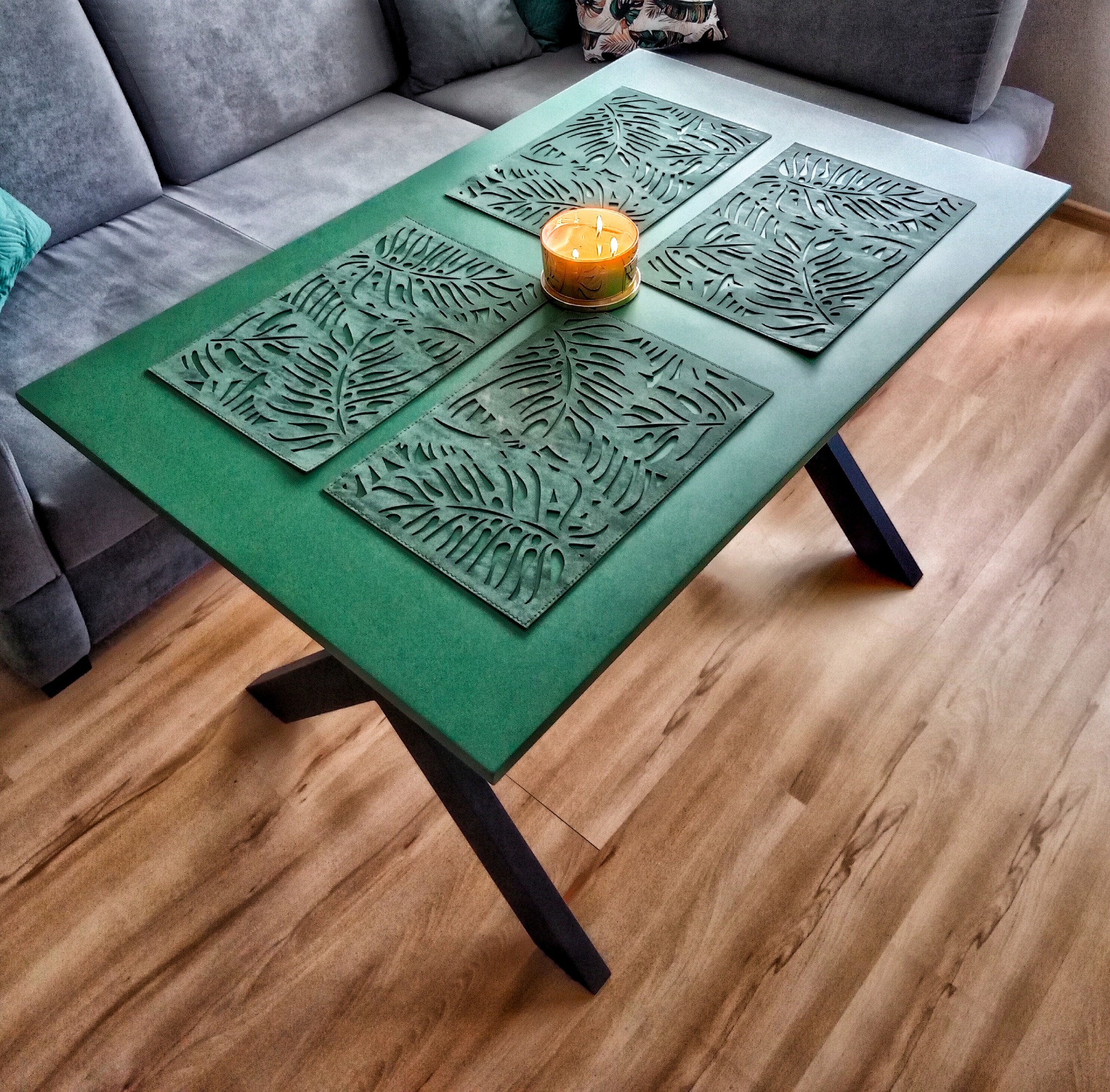 Stół z zielonym blatem i czarnymi nogami, na blacie cztery ażurowe podkładki z motywem liści i zapalona świeca, obok szara sofa.