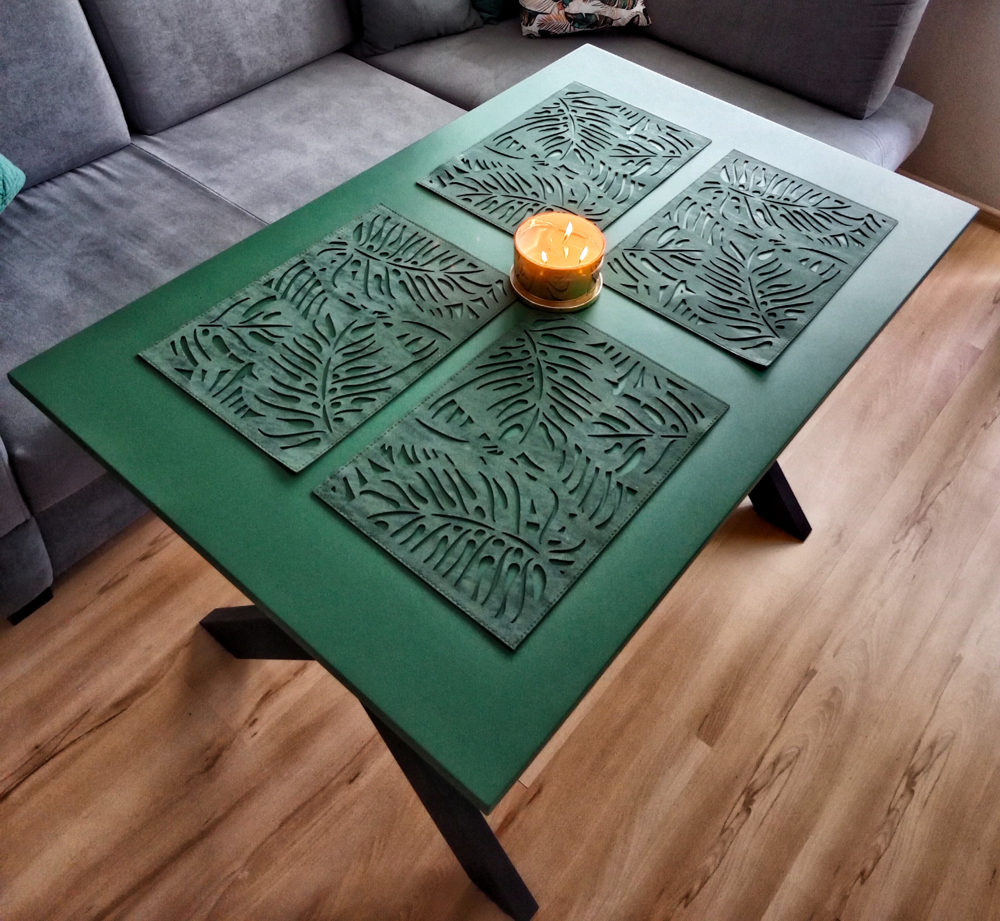 Zielony stolik kawowy z czterema ażurowymi podkładkami w kształcie liści i zapaloną świecą, na tle szarej kanapy i drewnianej podłogi.