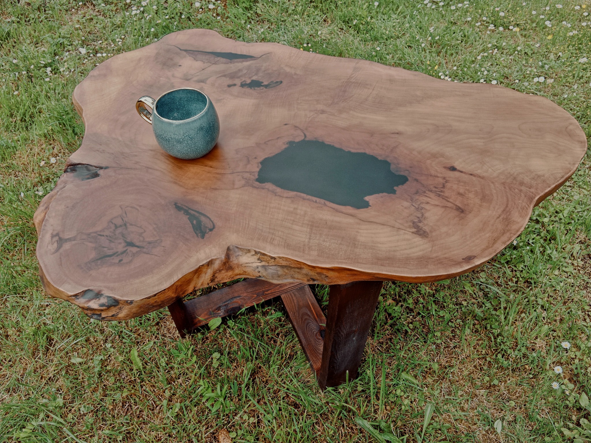 Stolik kawowy z blatem z naturalnego drewna z żywicą epoksydową, na trawie, z ceramicznym kubkiem z boku.