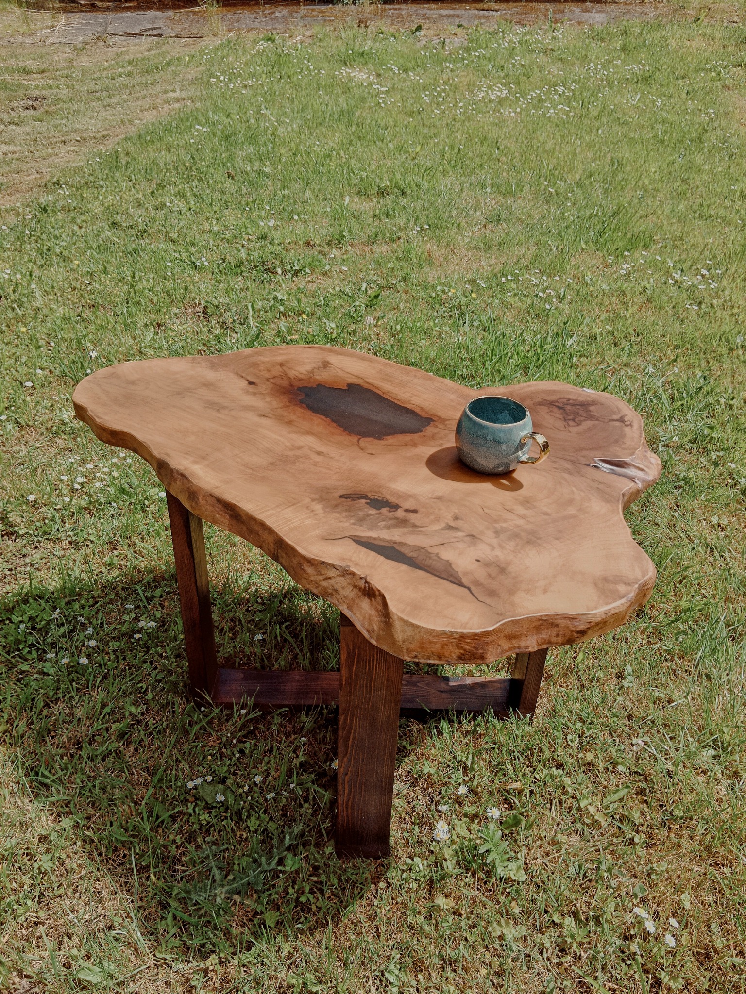 Stolik kawowy z blatem z naturalnego drewna o nieregularnym kształcie, z żywicą wypełniającą ubytki, stoi na trawie. Na blacie ceramiczny kubek z złotym uchem.