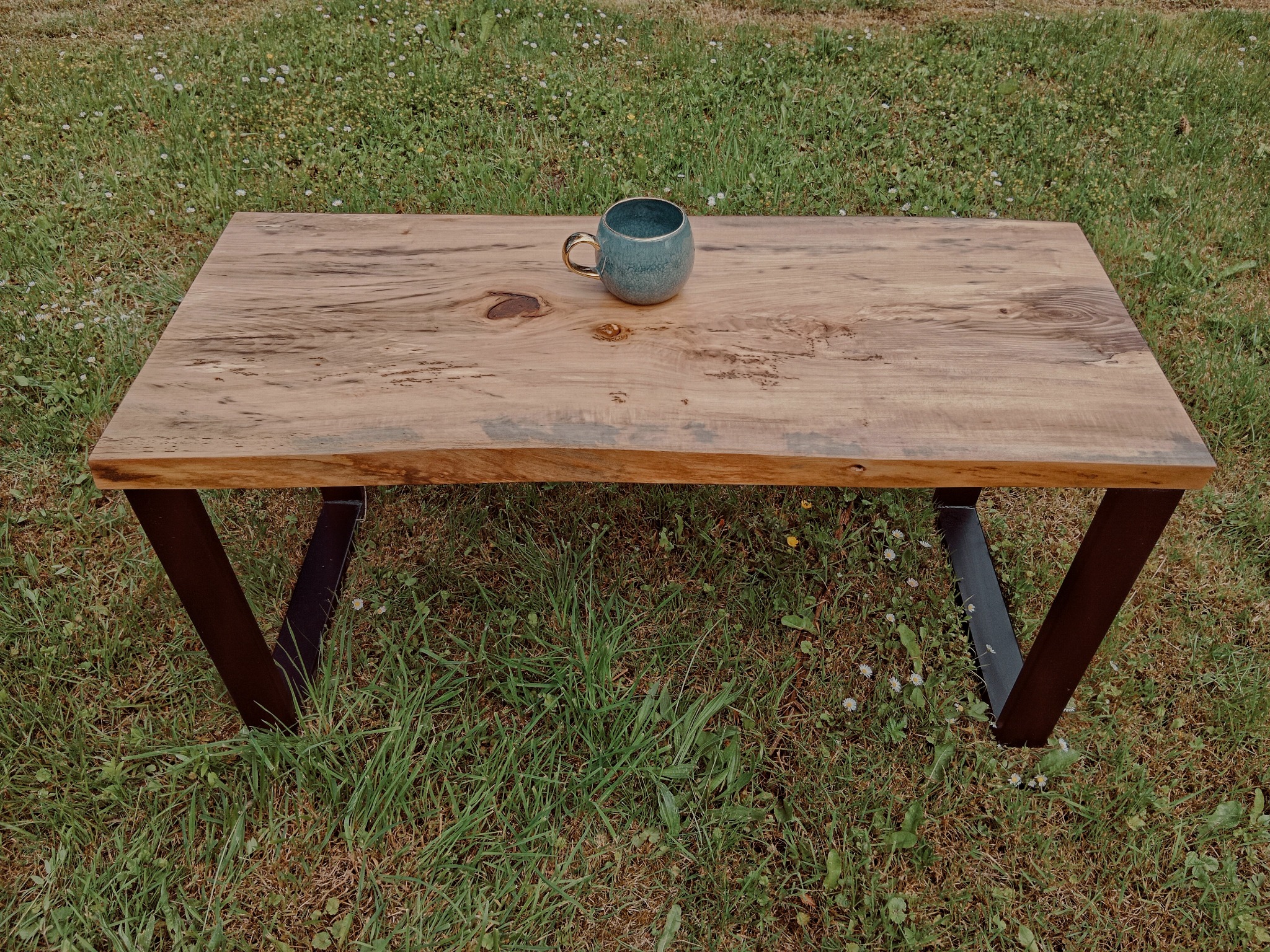 Niski stolik kawowy z blatem z jasnego drewna o nieregularnych krawędziach, wsparty na metalowych nogach w kształcie litery V, ustawiony na trawie, z ceramicznym kubkiem na blacie.
