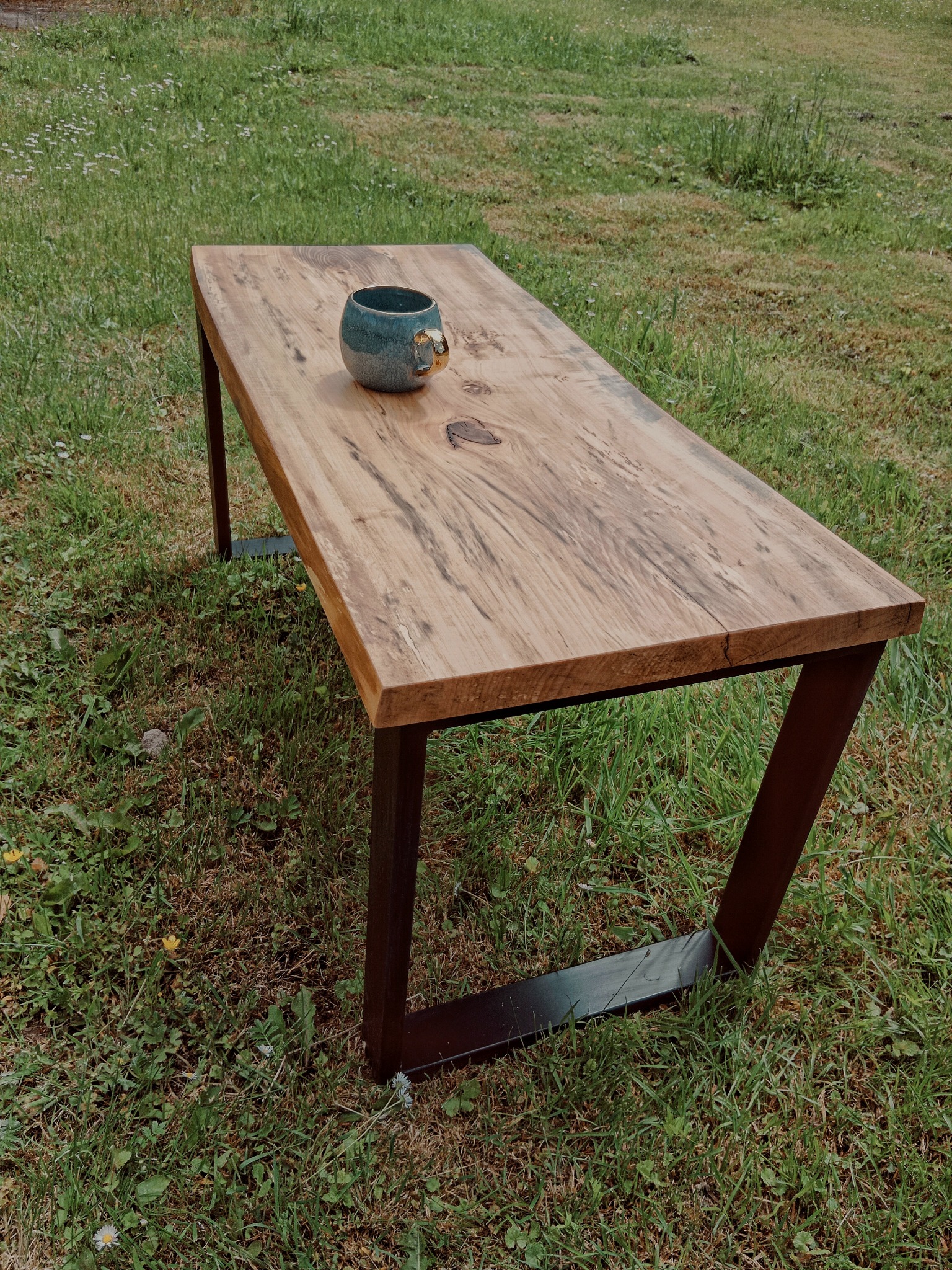 Drewniany stół na metalowych nogach, z widocznym usłojeniem drewna, ustawiony na trawie, z ceramicznym kubkiem na blacie.