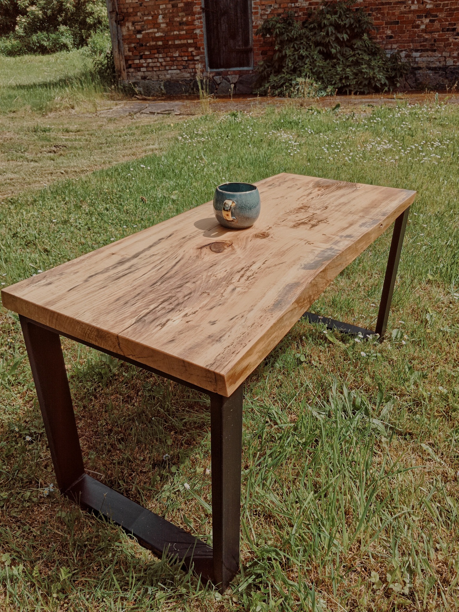 Stolik kawowy z blatem z jasnego drewna o wyraźnym usłojeniu, na metalowych nogach w kształcie litery U, ustawiony na trawie przed ceglanym budynkiem z ciemnymi drzwiami. Na blacie stoi ceramiczny...