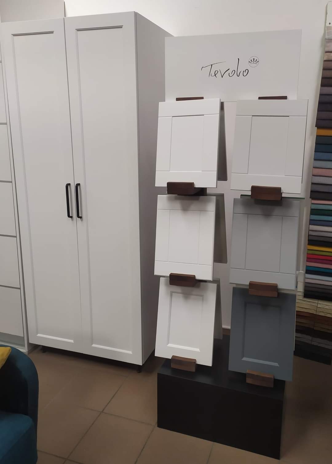 Ekspozycja próbek frontów szafek w różnych odcieniach bieli i szarości, prezentowanych obok białej, dwudrzwiowej szafy z minimalistycznymi, czarnymi uchwytami.