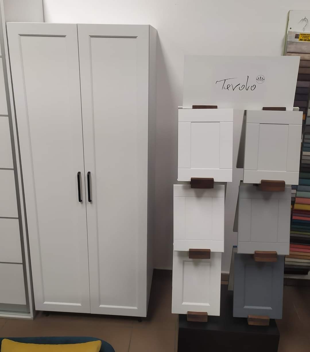 Biała, dwudrzwiowa szafa z czarnymi uchwytami, obok ekspozycja próbek frontów meblowych w różnych odcieniach szarości i bieli z drewnianymi uchwytami, sygnowanych logo 'Tevolo'.