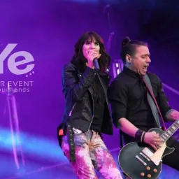 Zespół muzyczny na scenie podczas występu: wokalistka z mikrofonem w skórzanej kurtce i gitarzysta z czarną gitarą elektryczną, fioletowe oświetlenie sceniczne i logo firmy eventowej w tle.
