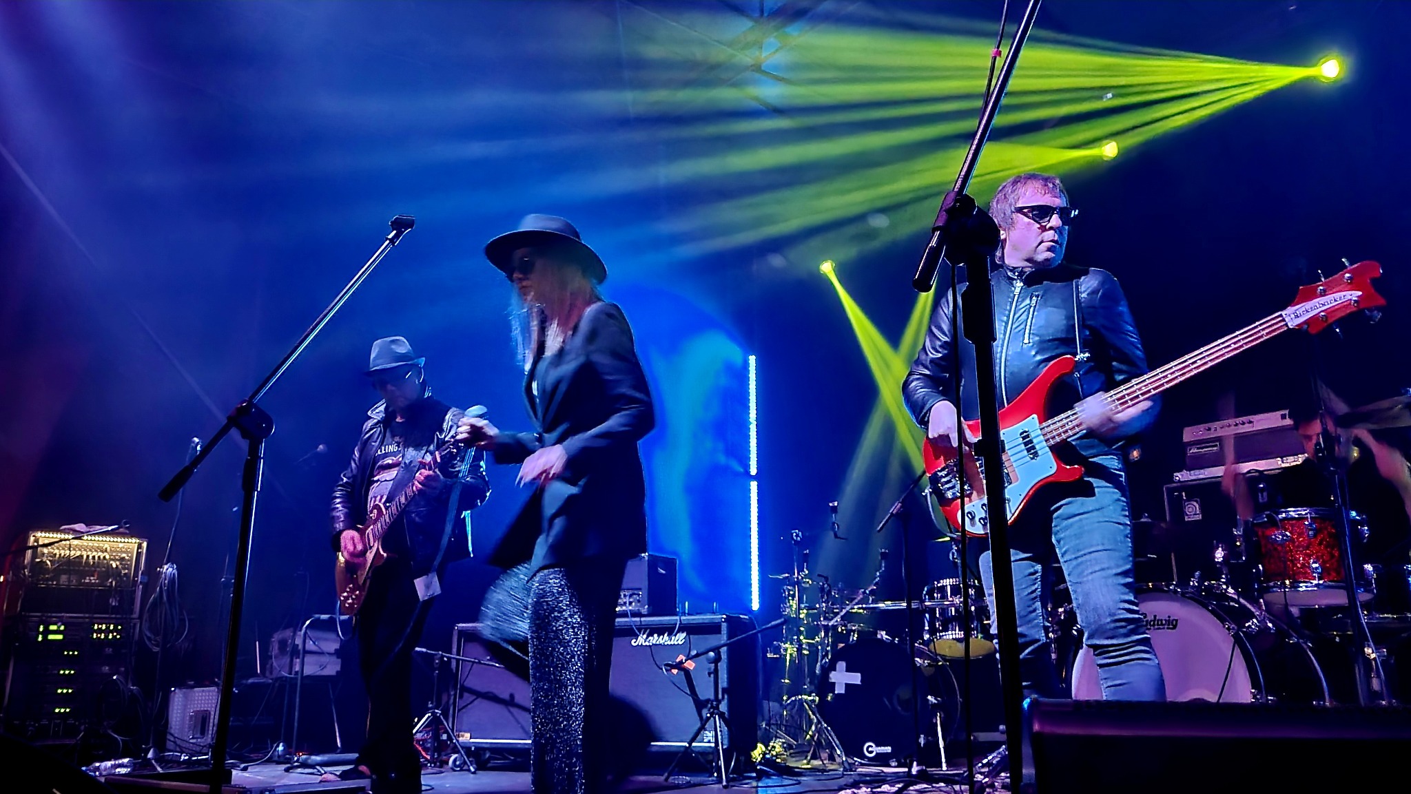 Zespół muzyczny na scenie podczas występu; kobieta w czarnym kapeluszu i błyszczących spodniach, gitarzysta w skórzanej kurtce i basista z czerwoną gitarą basową Rickenbacker, oświetlenie sceniczne...