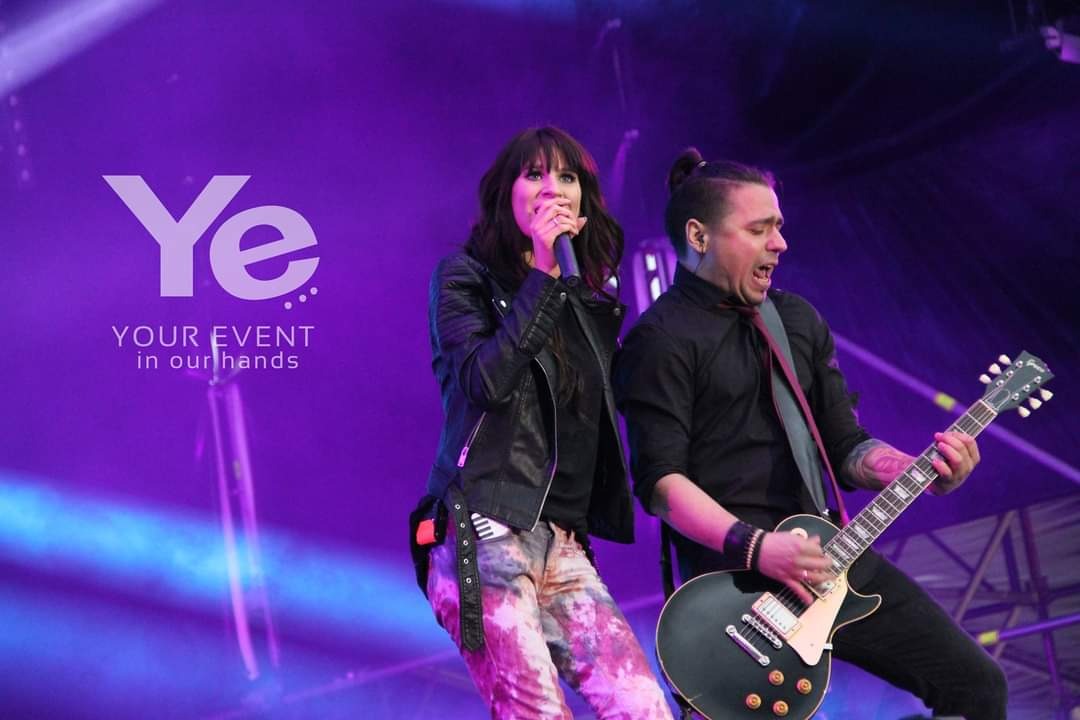 Zespół muzyczny na scenie podczas występu: wokalistka z mikrofonem w skórzanej kurtce i gitarzysta z czarną gitarą elektryczną, fioletowe oświetlenie sceniczne i logo firmy eventowej w tle.